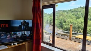 A room with a TV displaying a Netflix interface, a wooden deck with a railing, and a view of a forested hillside. - The Meetings B&B (Avoca)