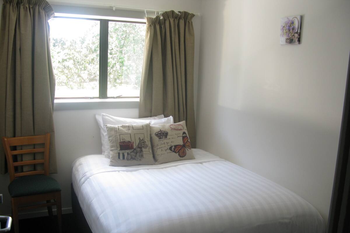 Un dormitorio con cama, dos almohadas, una silla y una ventana con cortinas.