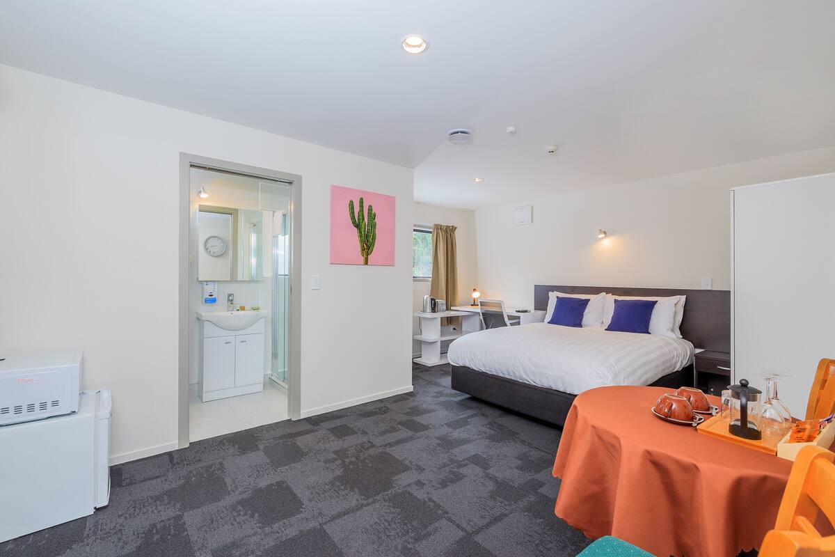Habitación de hotel con cama, escritorio, silla, mesa con bandeja, lavamanos y espejo.