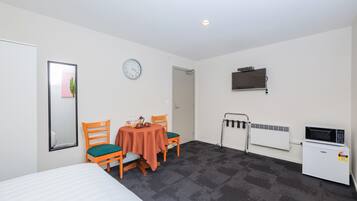 Una habitación con cama, una mesa con sillas, un reloj, un televisor, un radiador y una puerta.