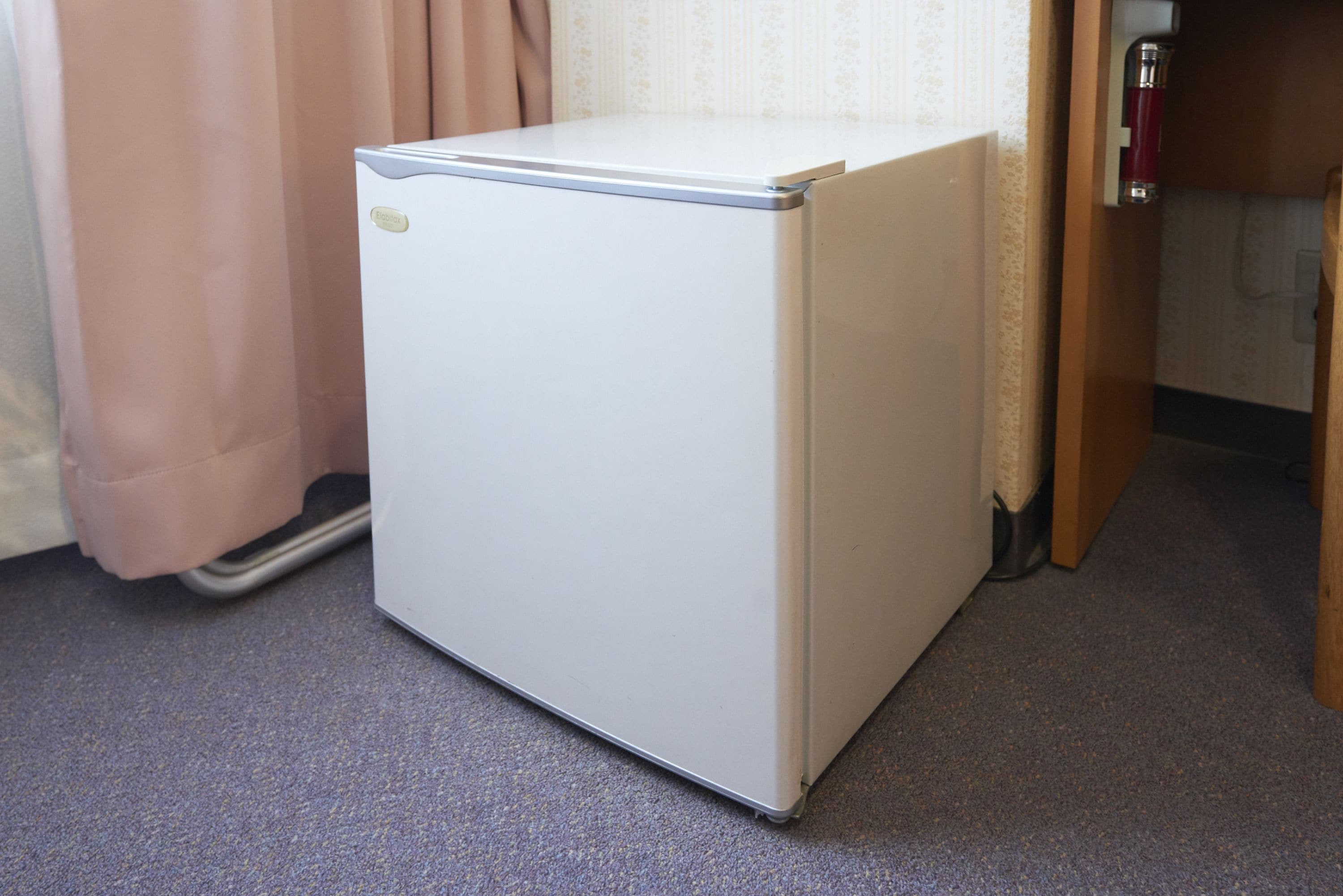 Un refrigerador pequeño y blanco en un rincón de una habitación con piso alfombrado y una puerta de madera.