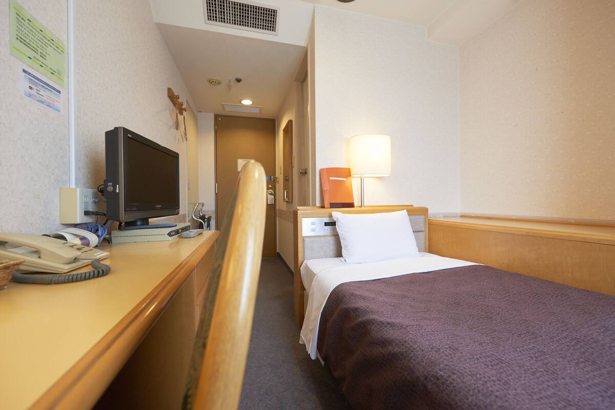 A hotel room with a bed, bedside table, lamp, and a desk with a computer and phone.