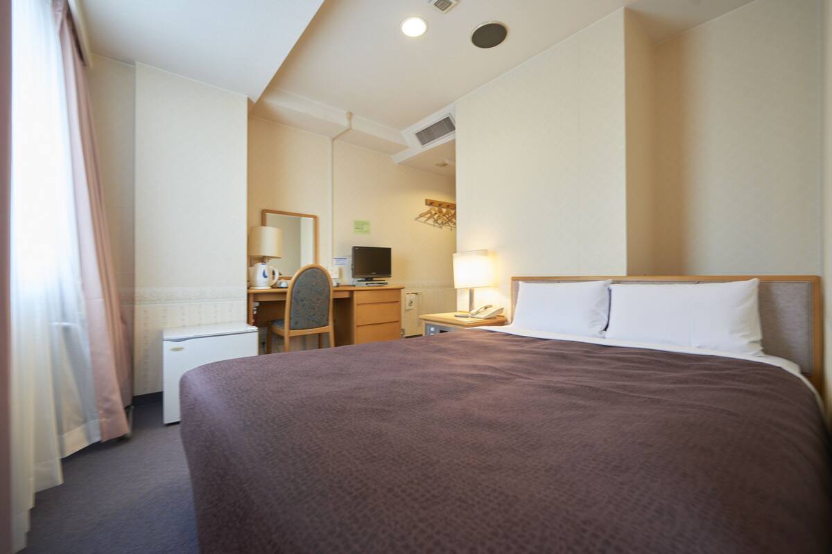 A hotel room with a bed, a desk, a chair, a small refrigerator, and a lamp.