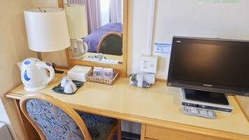 Quarto de hotel com secretária de madeira, televisão, candeeiro, espelho e cama com manta roxa.