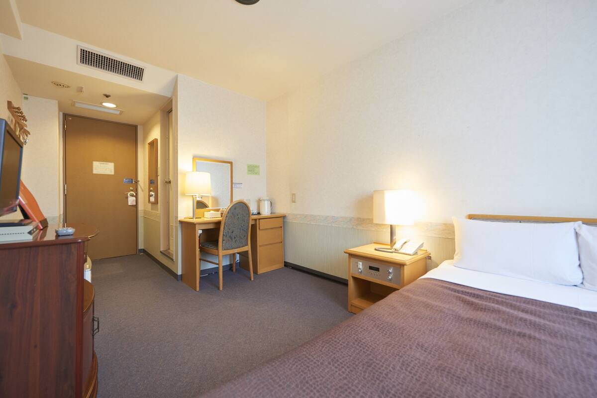 A hotel room with a bed, desk, chair, and a small bedside table with a lamp.
