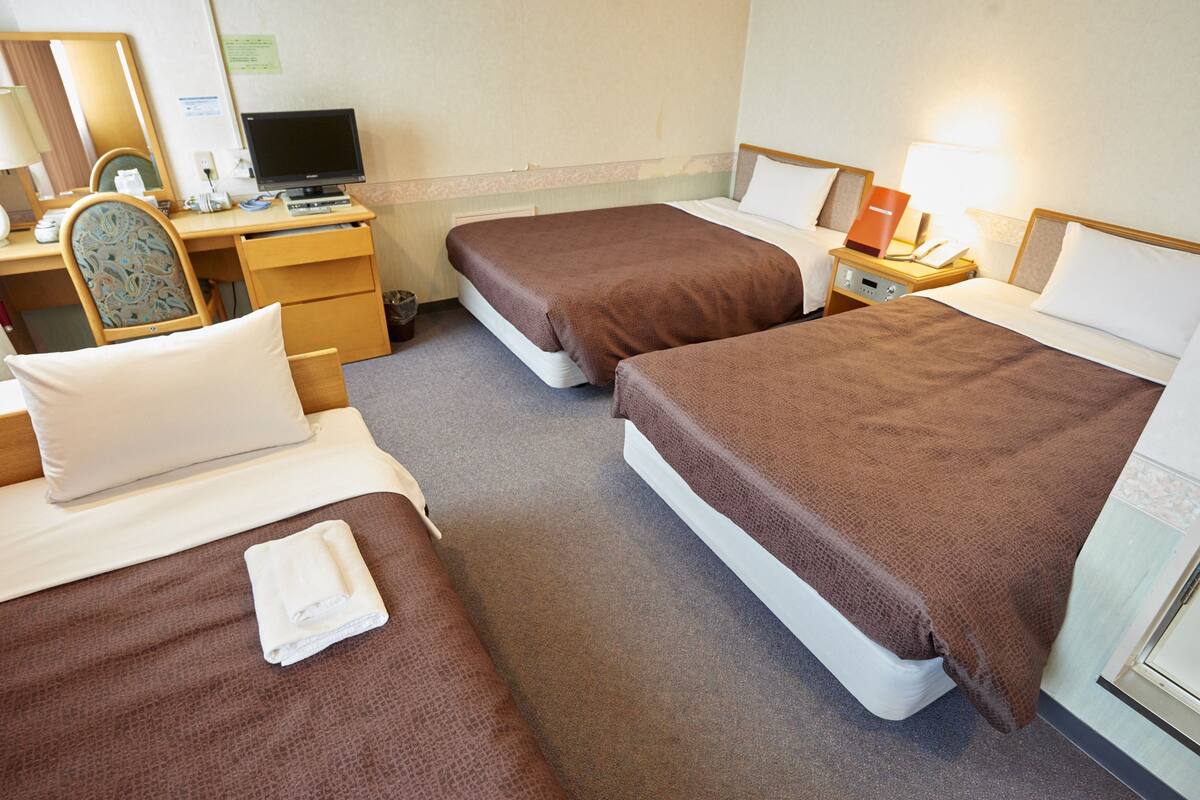A hotel room with two beds, a desk with a computer, a chair, and a small table with a lamp.