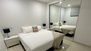 A modern bedroom with a large bed, a nightstand with a lamp, a bedside table, and a mirror. - Magnum Serviced Apartments (Darwin)