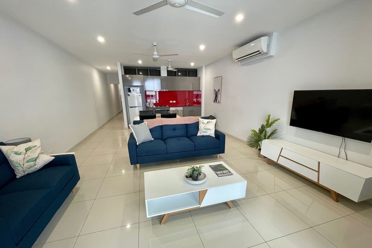 A modern living room with a blue sofa, a white coffee table with a plant, a flat-screen TV, and a kitchen in the background.