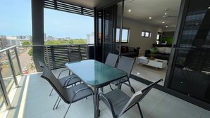 A modern balcony with a glass table and chairs, a sofa, and a kitchen area visible through the glass doors. - Magnum Serviced Apartments (Darwin)