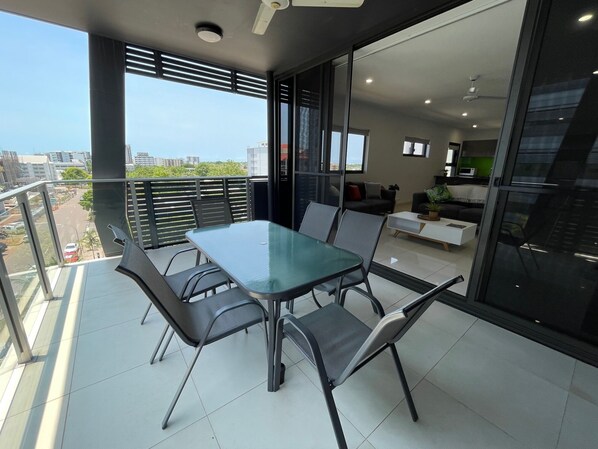 A modern balcony with a glass table and chairs, a sofa, and a kitchen area visible through the glass doors. - Magnum Serviced Apartments (Darwin)