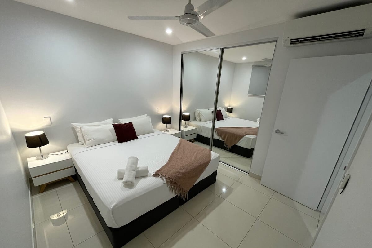 A modern bedroom with a large bed, bedside tables with lamps, a ceiling fan, and a mirrored closet door.