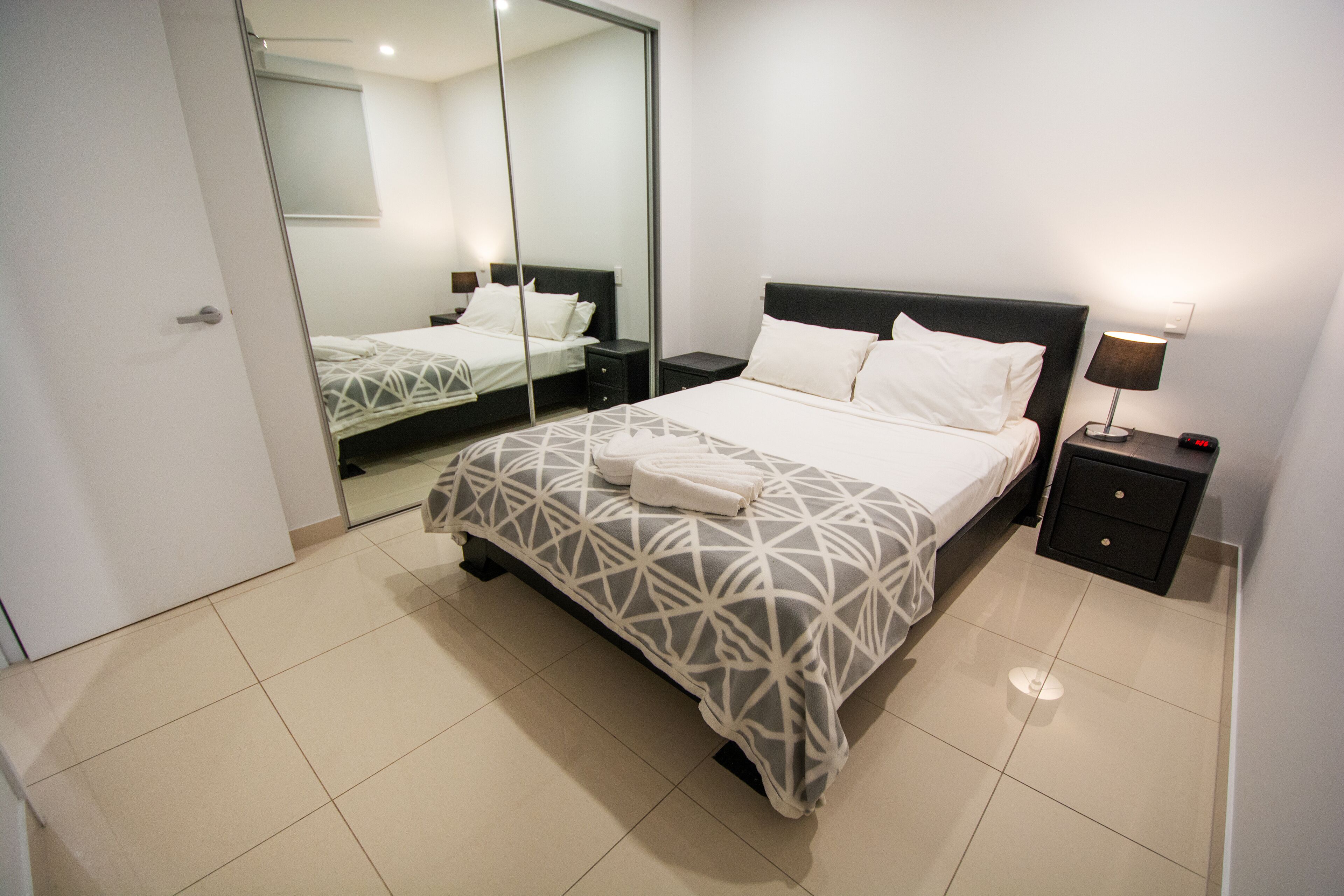 A modern hotel room with a large bed, a bedside table with a lamp, and a mirror on the wall.