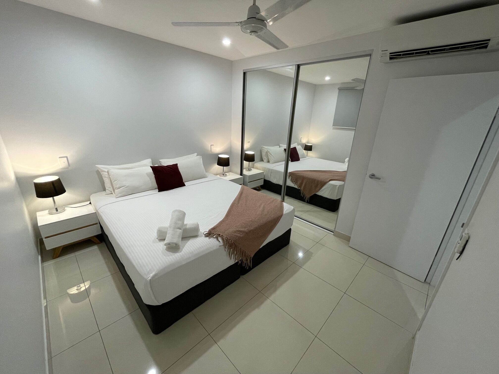 A modern bedroom with a large bed, a bedside table with a lamp, a ceiling fan, and a mirror on the wall.