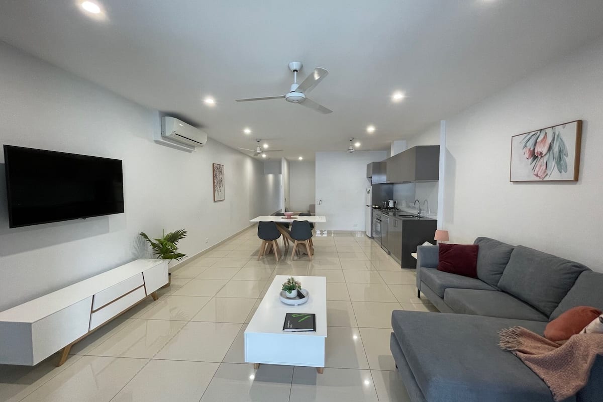 A modern living room with a flat-screen TV, a sofa, a coffee table, and a dining area with chairs and a table.