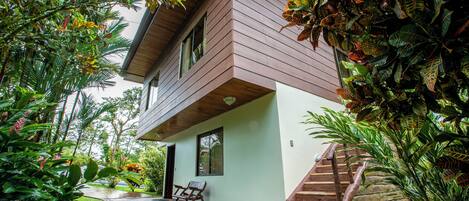 A two-story house with a covered porch, a wooden staircase, and lush tropical plants.