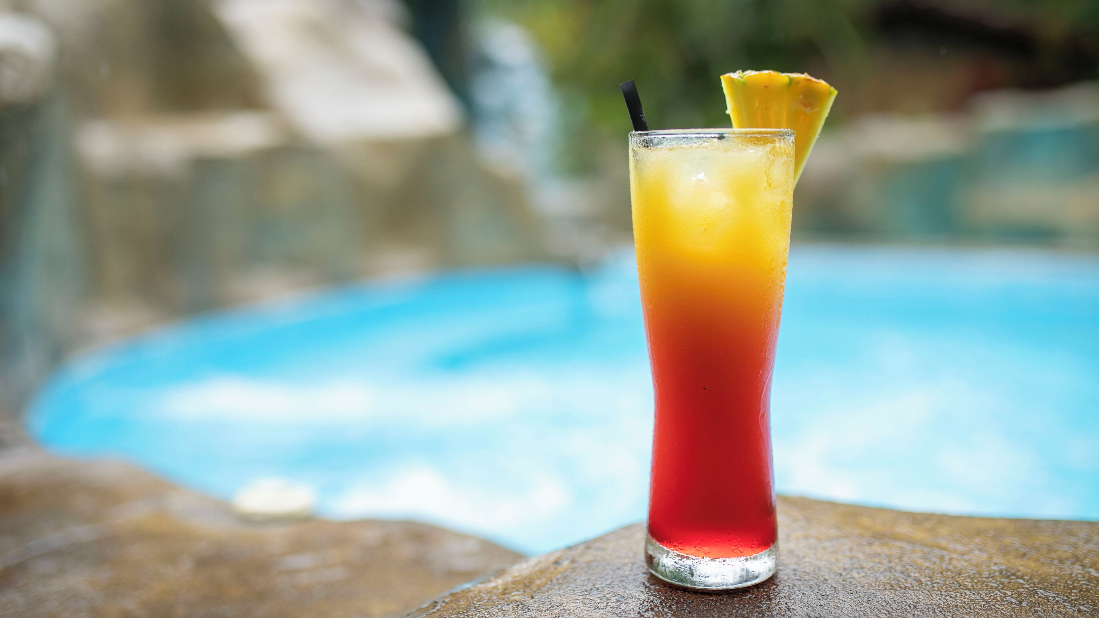 Un vaso alto con una bebida degradada, una pajita y una rodaja de piña, sobre una superficie de piedra junto a una piscina.