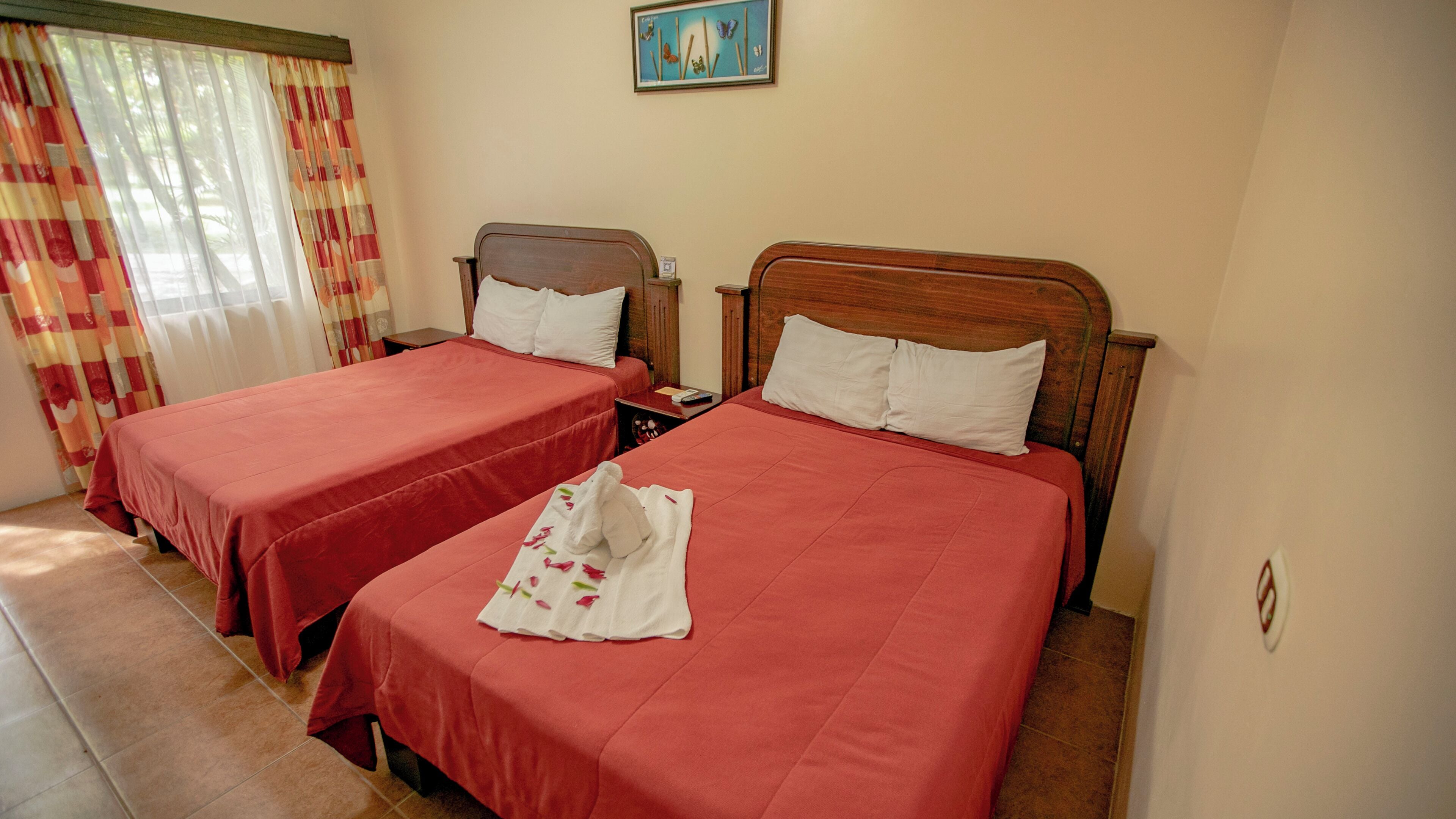 Ein Hotelzimmer mit zwei Betten, jeweils mit einem hölzernen Kopfteil, roter Bettwäsche und einem weißen Handtuch mit floralem Muster auf einem der Betten.