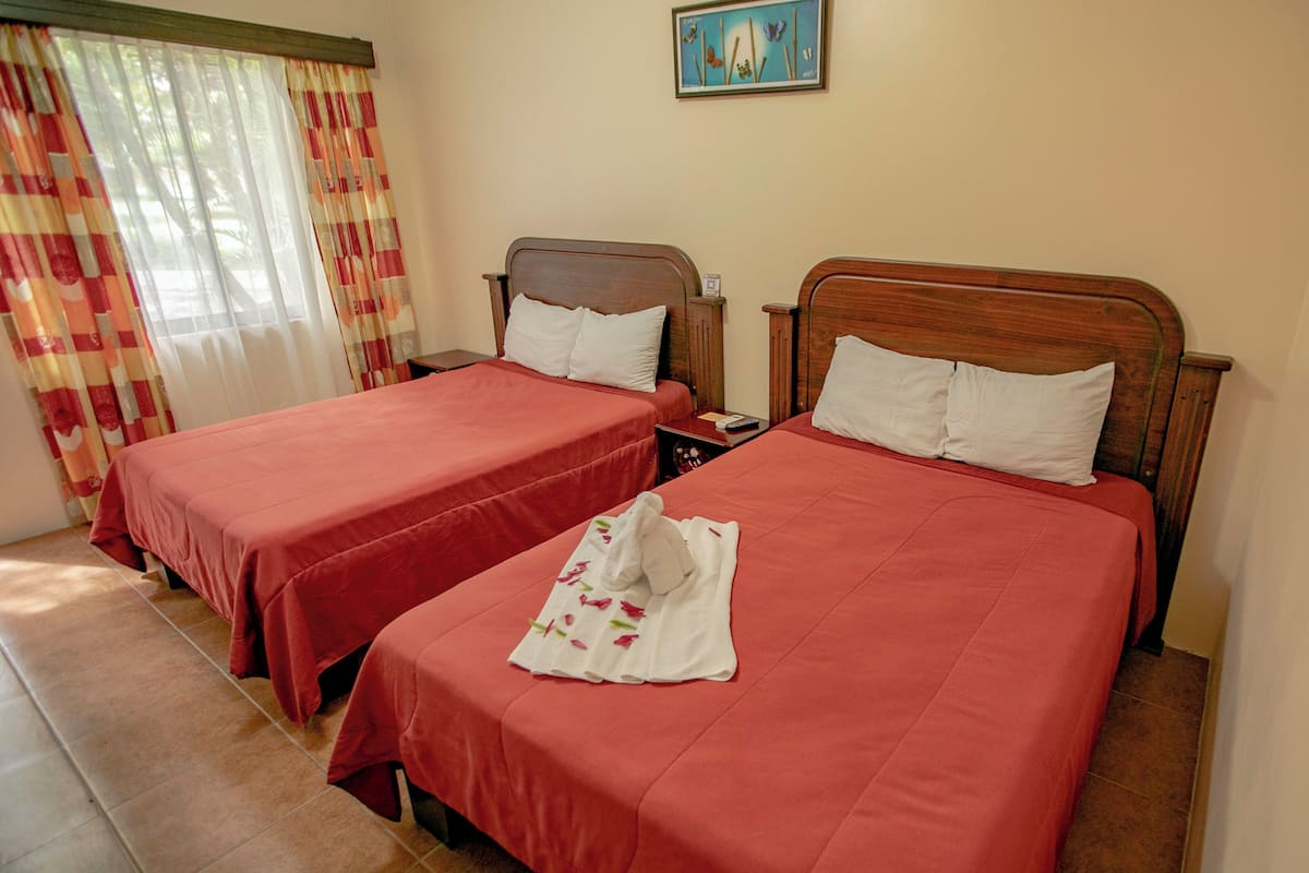 A hotel room with two beds, each with a wooden headboard, red bedding, and a white towel with floral patterns on one bed.