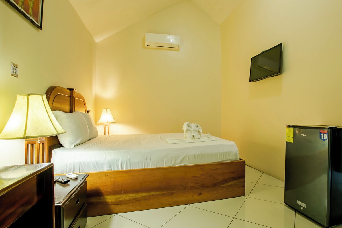 Una habitación de hotel con cama, mesita de noche, lámpara, televisor y refrigerador.