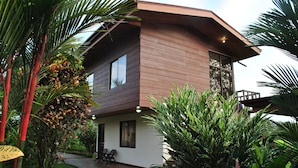 A two-story house with a brown exterior, a balcony, and a small porch with chairs. - Hotel Las Orquideas (La Fortuna)