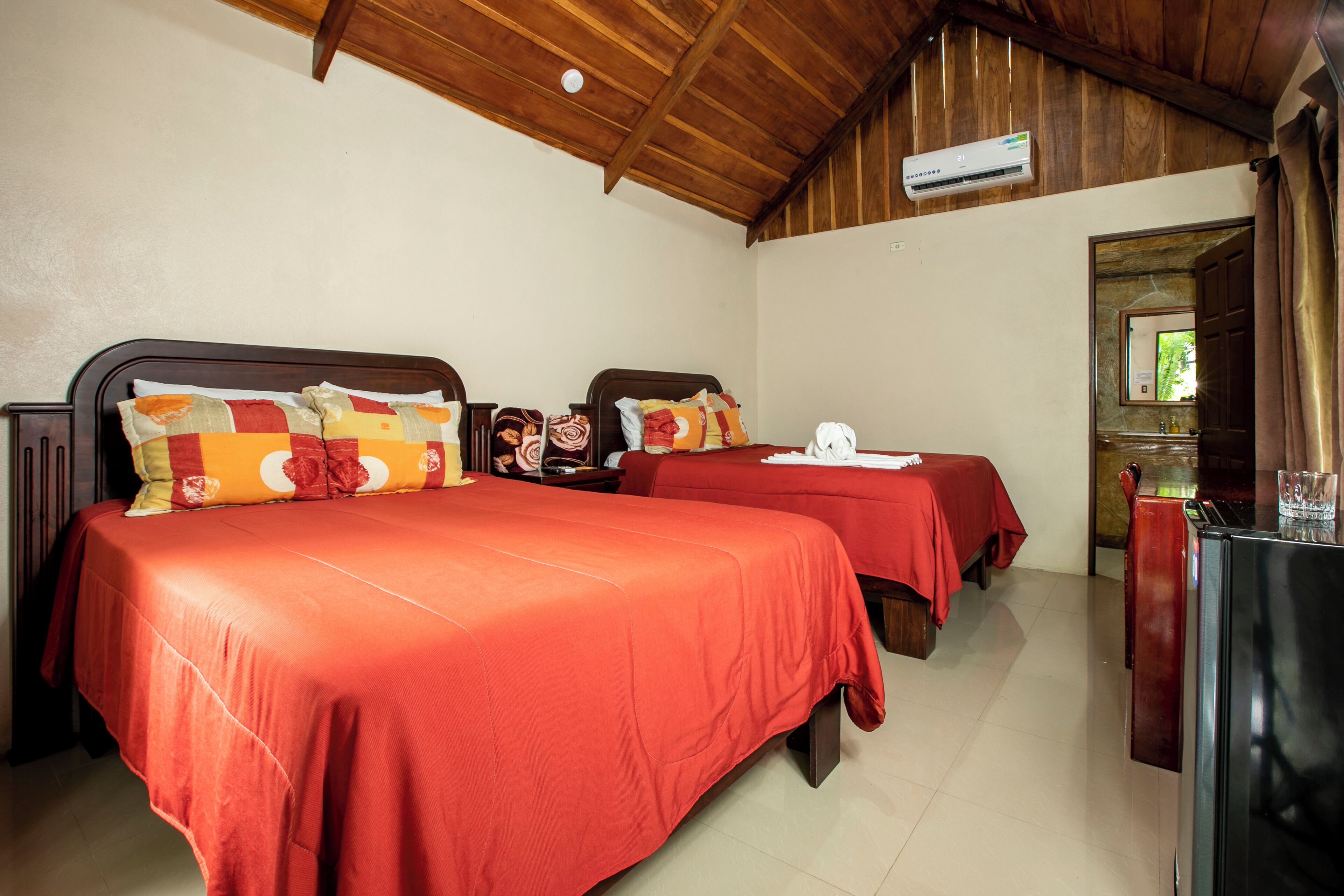 A room with two beds, each with red bedding and decorative pillows. There is a wooden ceiling and an air conditioning unit mounted on it.