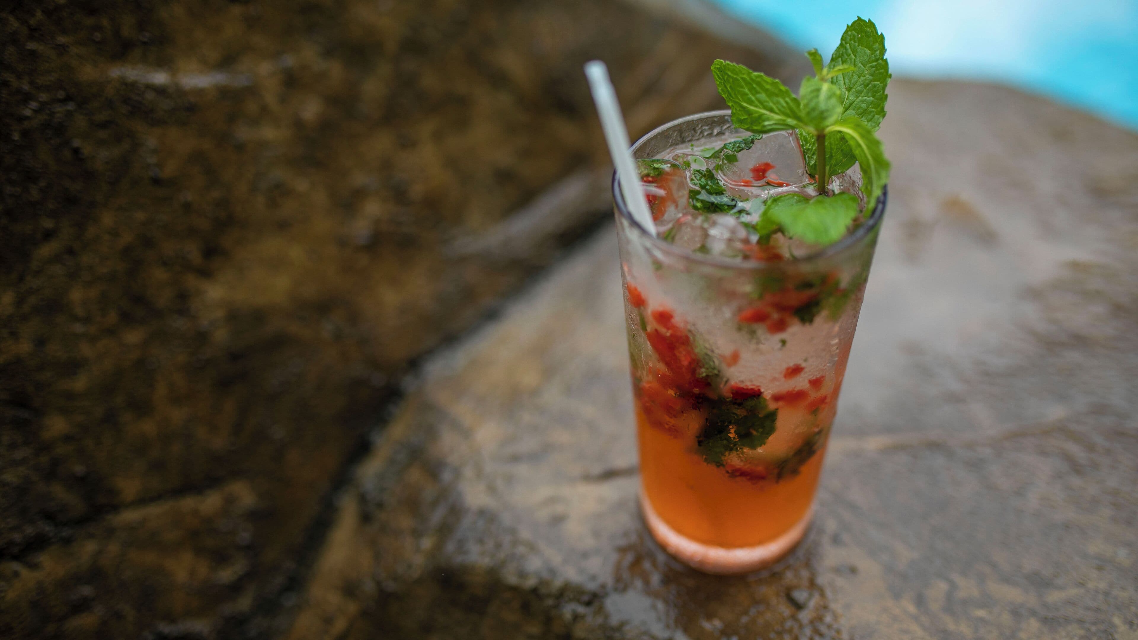 A glass of iced drink with mint and fruit on a stone surface.