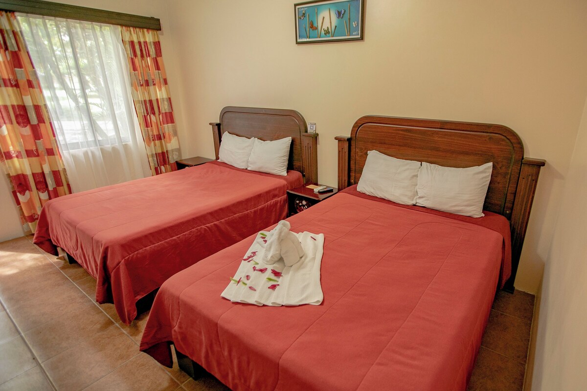 Habitación de hotel con dos camas, cada una con cabecero de madera, ropa de cama roja y una toalla blanca con motivos florales sobre una de ellas.