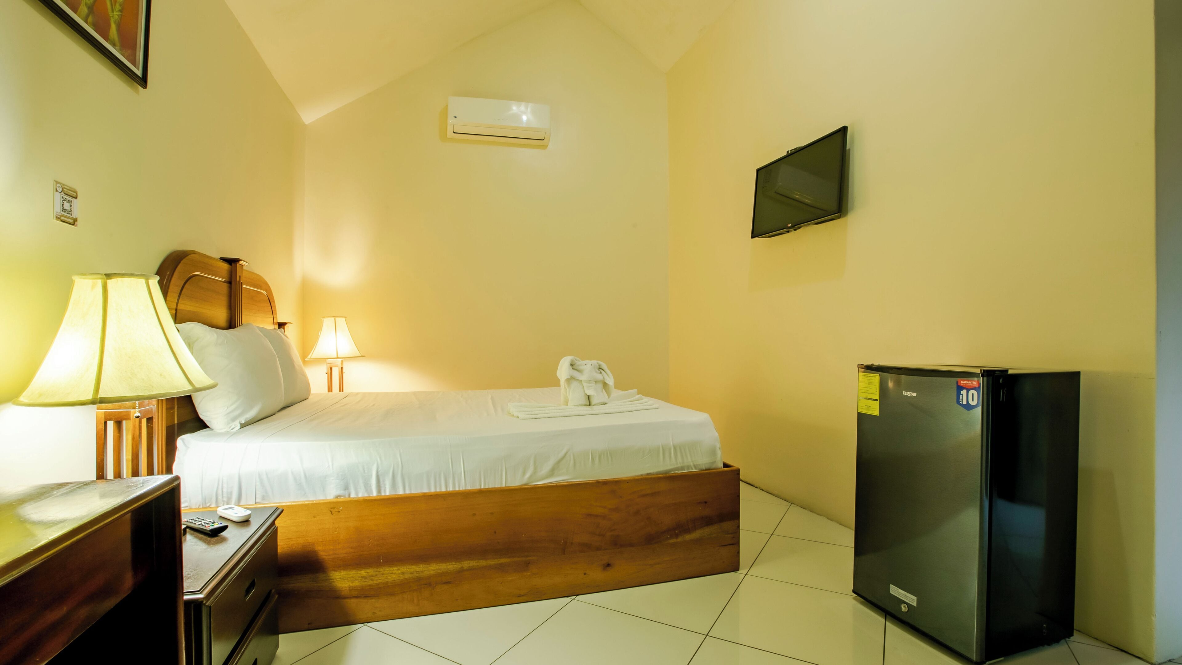 A hotel room with a bed, a nightstand, a lamp, a TV, and a refrigerator.