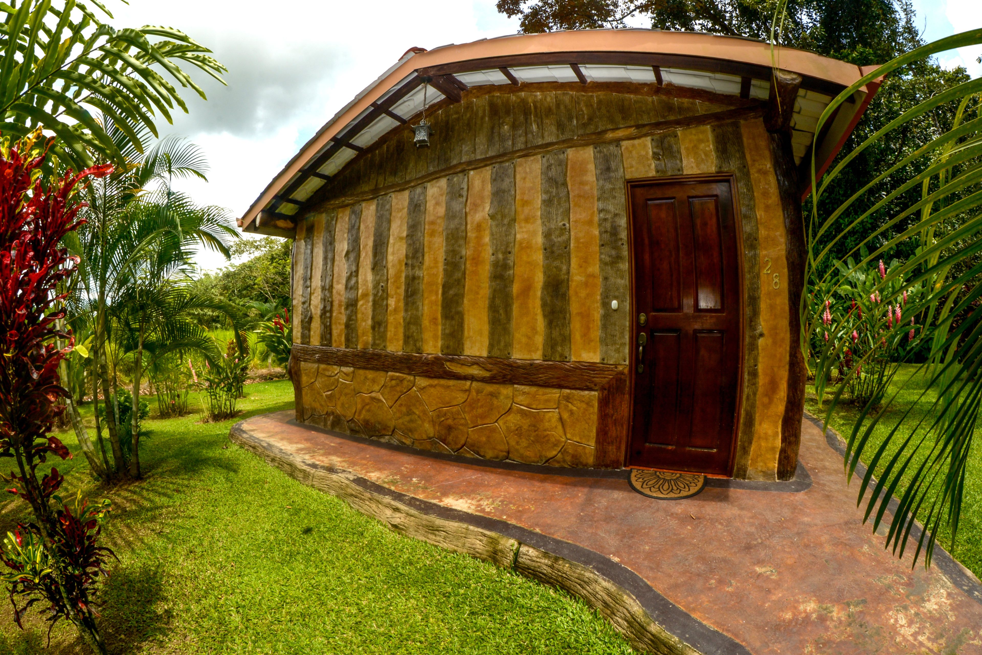 A rustic wooden building with a curved roof and a wooden door, surrounded by lush greenery.