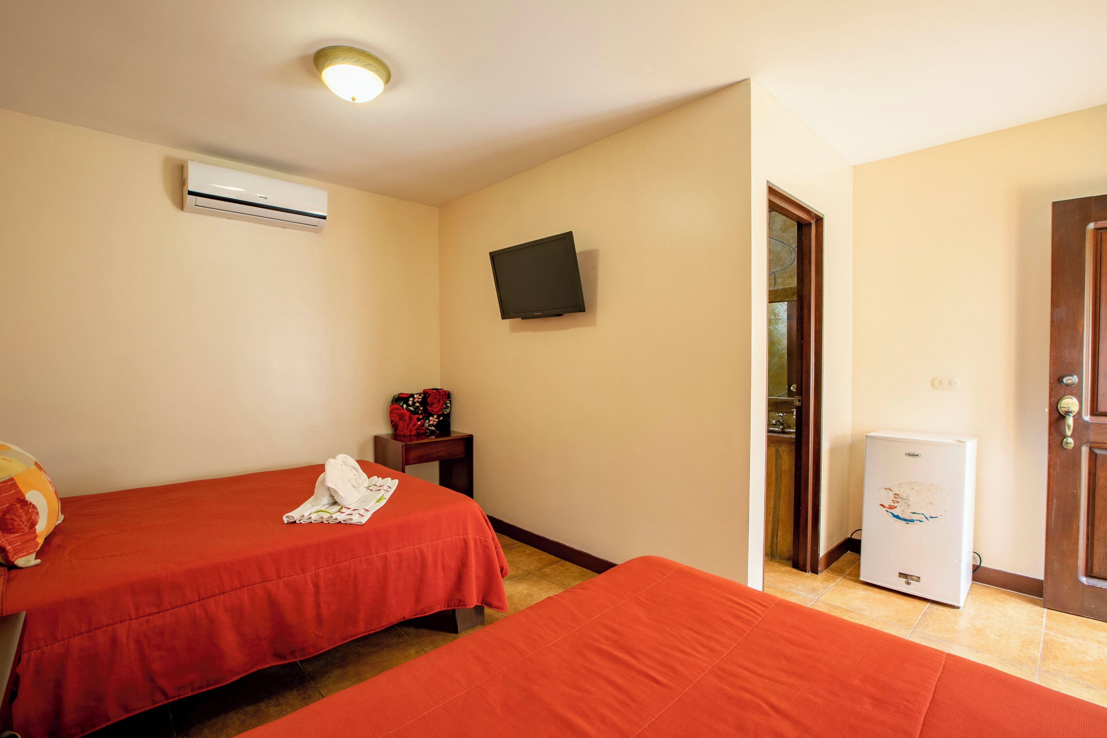 Ein Hotelzimmer mit zwei Betten, einem Fernseher, einer Klimaanlage, einem Kühlschrank und einer Tür.