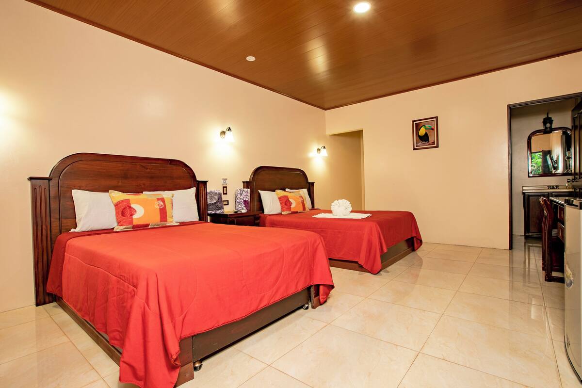 A hotel room with two beds, each with a red bedspread and white pillows. There are wall-mounted lights above each bed, and a small framed picture on the wall.