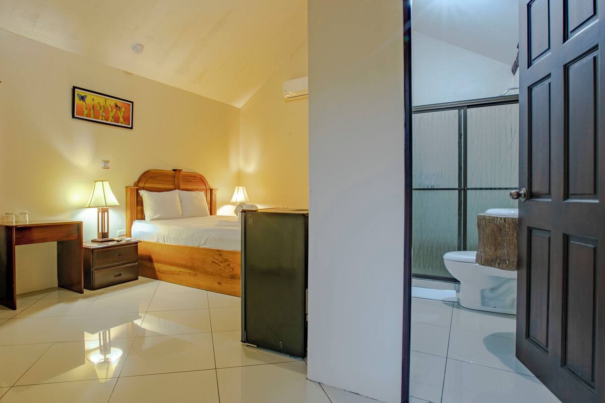 A hotel room with a bed, bedside tables, a nightstand, a lamp, a picture on the wall, and a view of the bathroom through an open door.