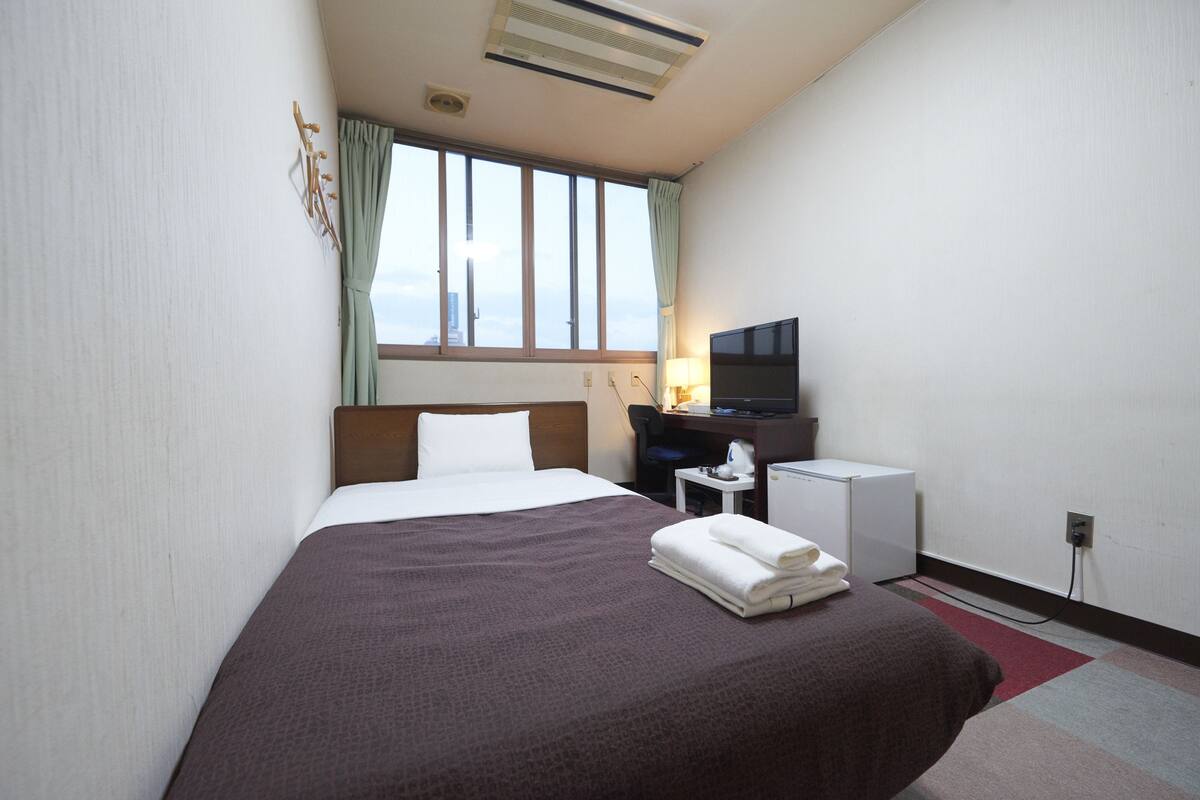 Quarto de hotel com cama, escrivaninha, televisão e janela com cortinas.