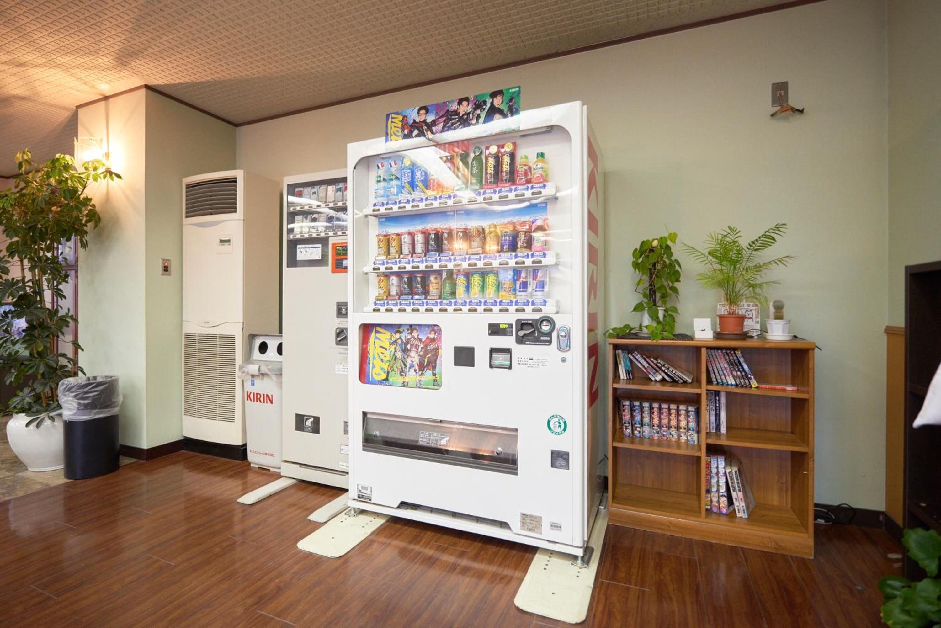 A vending machine with various drinks and snacks, a trash bin, a potted plant, and a bookshelf with magazines.