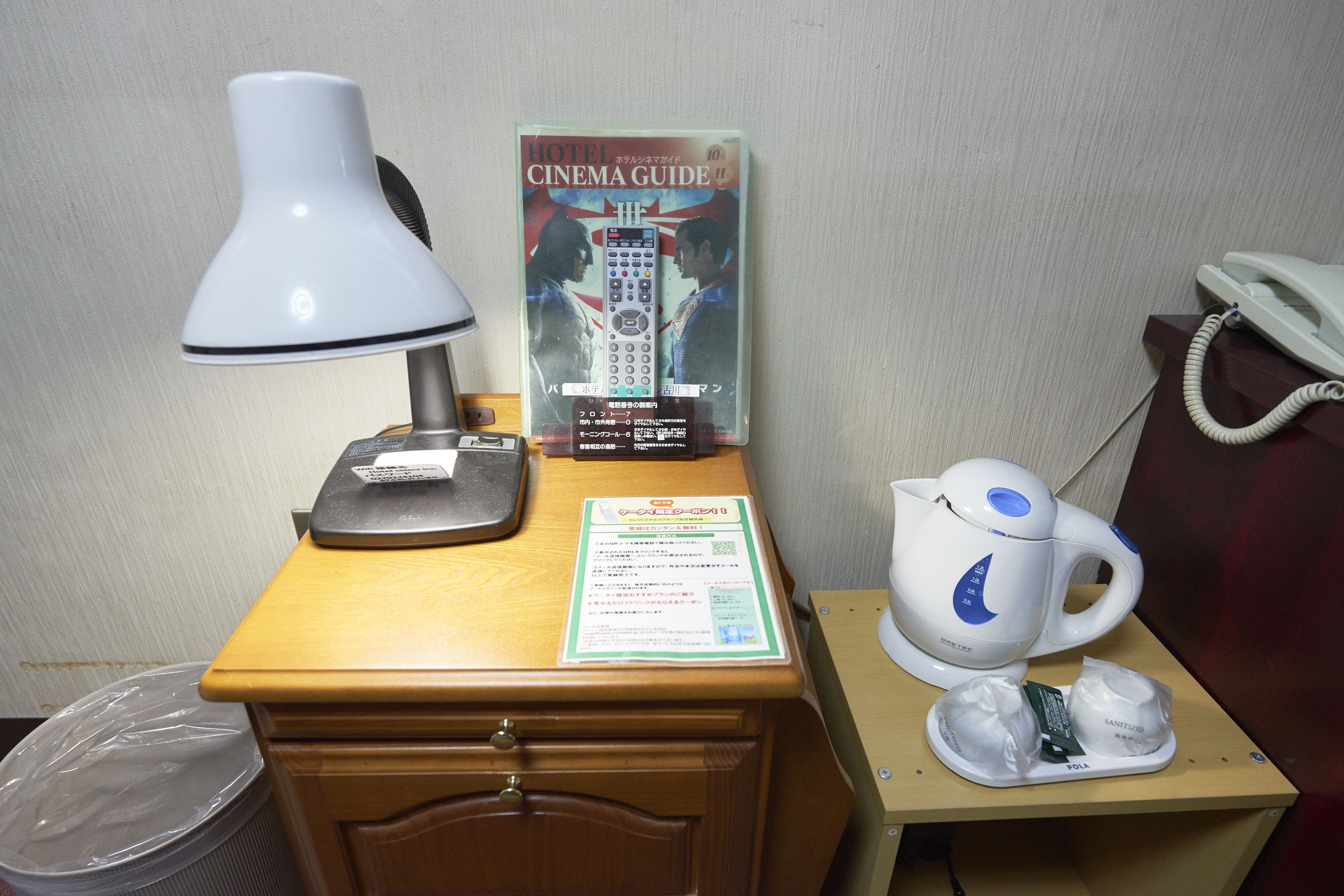 A hotel room desk with a lamp, a cinema guide, a phone, a kettle, and a trash bin.