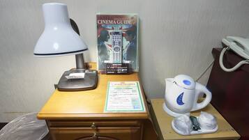 A hotel room desk with a lamp, a cinema guide, a phone, a kettle, and a trash bin.