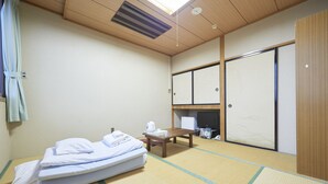Ein Zimmer mit Tatami-Boden, einem Bett, einem Holztisch, einem Fernseher und Schiebetüren.