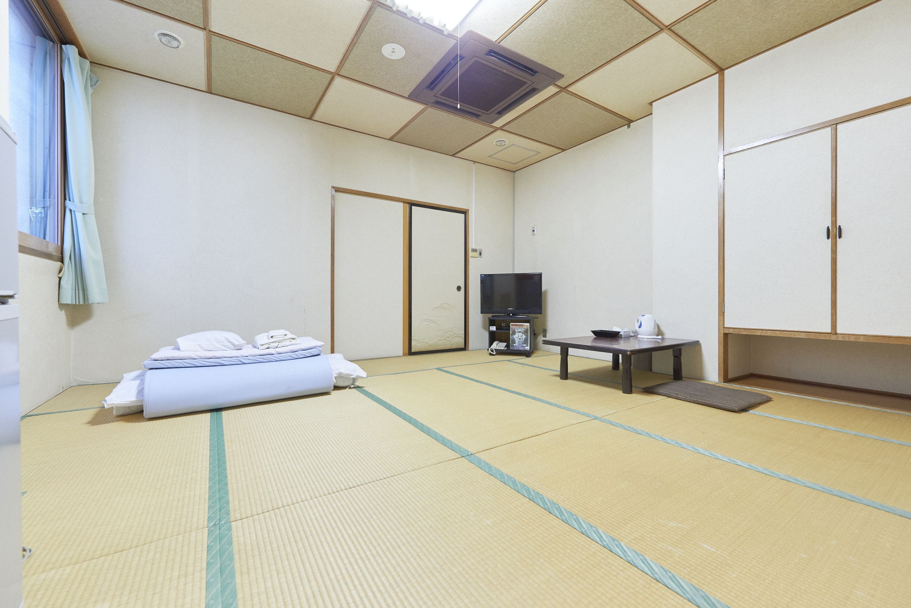Una habitación japonesa tradicional con suelo de tatami, una cama con ropa de cama blanca, un televisor, una mesita con un hervidor y puertas correderas.