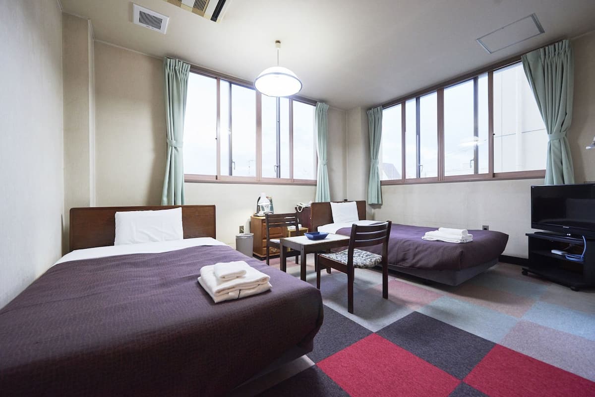 Quarto de hotel com duas camas, uma escrivaninha, uma cadeira, televisão e janelas amplas com cortinas.