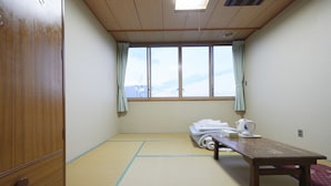 Ein Raum mit Tatami-Matten, einem Holztisch, einem Fenster mit Vorhängen und einem Holzschrank.
