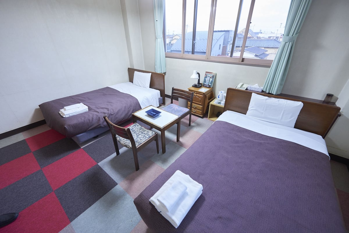 Quarto de hotel com duas camas, uma escrivaninha, uma cadeira, uma mesinha de cabeceira e janela com vista para edifícios.
