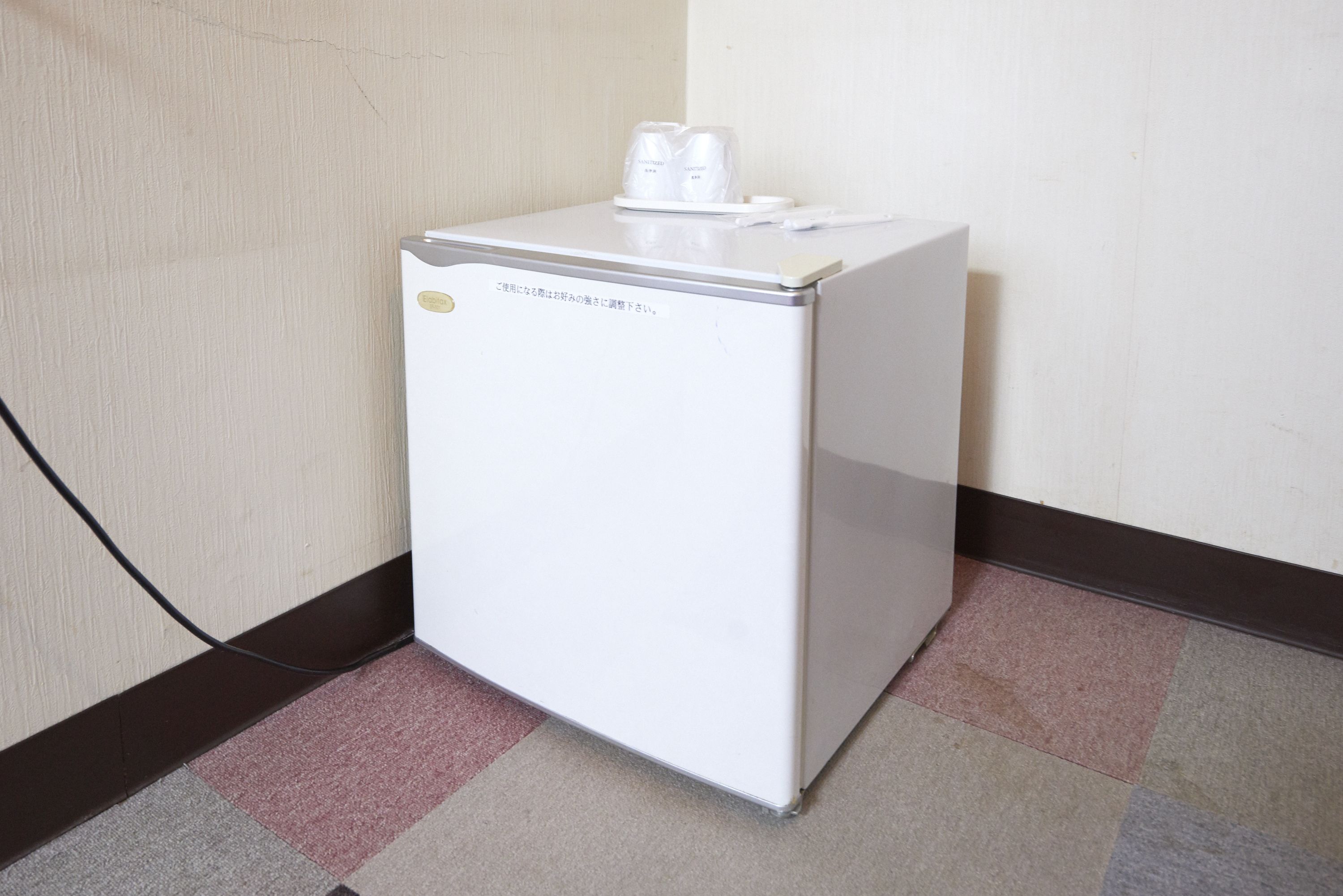 A small white refrigerator with a water dispenser and a coffee maker on top, located in a corner with a patterned carpet and a plain wall.