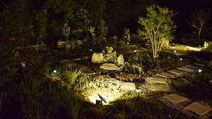 Un jardin nocturne avec des allées pavées de pierres, éclairé d’une lumière douce, et entouré d’un feuillage et d’arbres luxuriants.