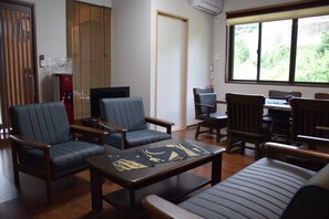 A room with a sofa, two armchairs, a coffee table, and a dining table with chairs. There is a window with a view of greenery outside.