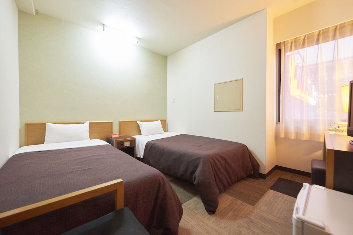 A hotel room with two single beds, a desk, a chair, a small table, and a window with curtains.