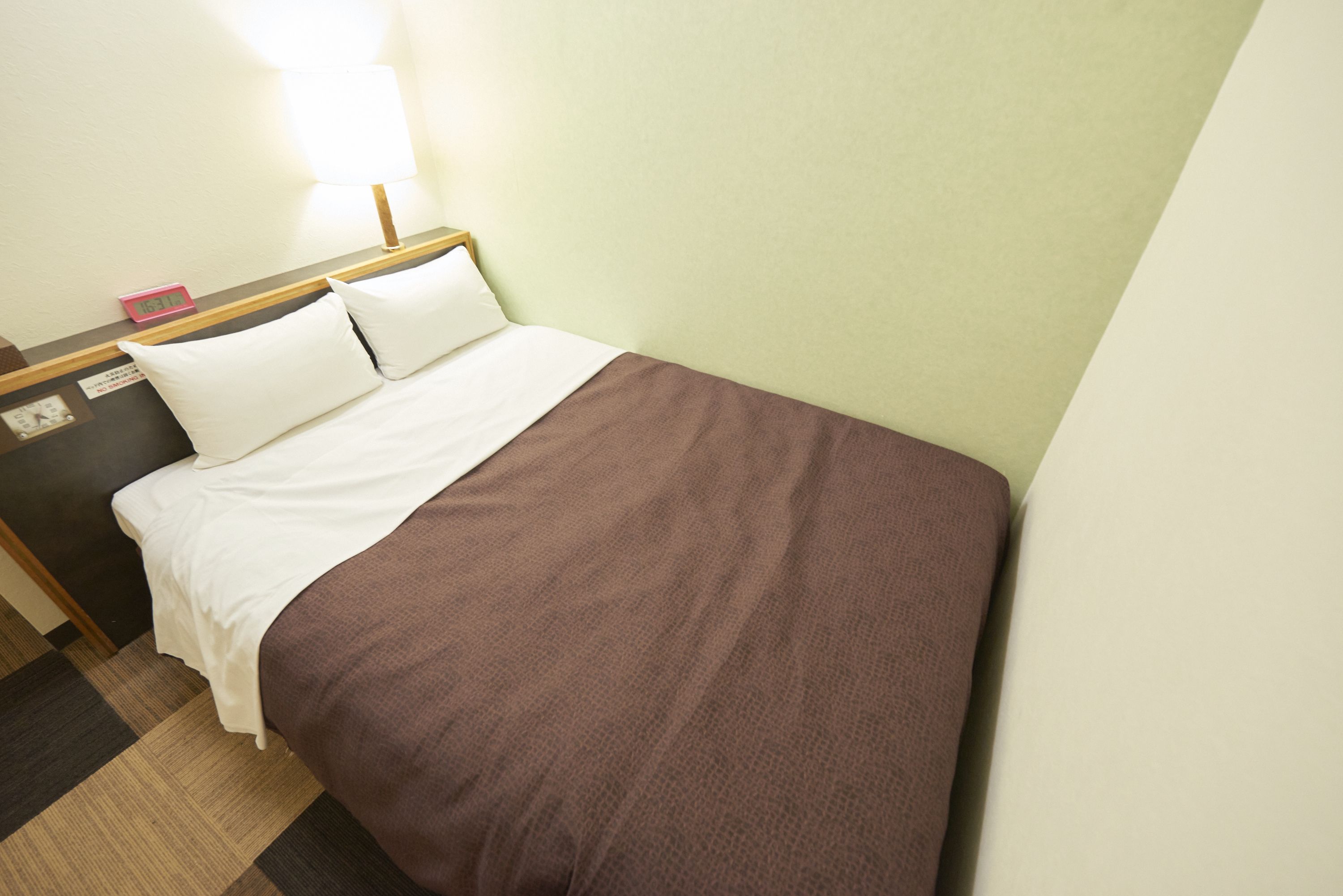 Un letto rifatto con cura, con biancheria bianca e marrone, una testiera in legno e un comodino con una lampada e una sveglia.