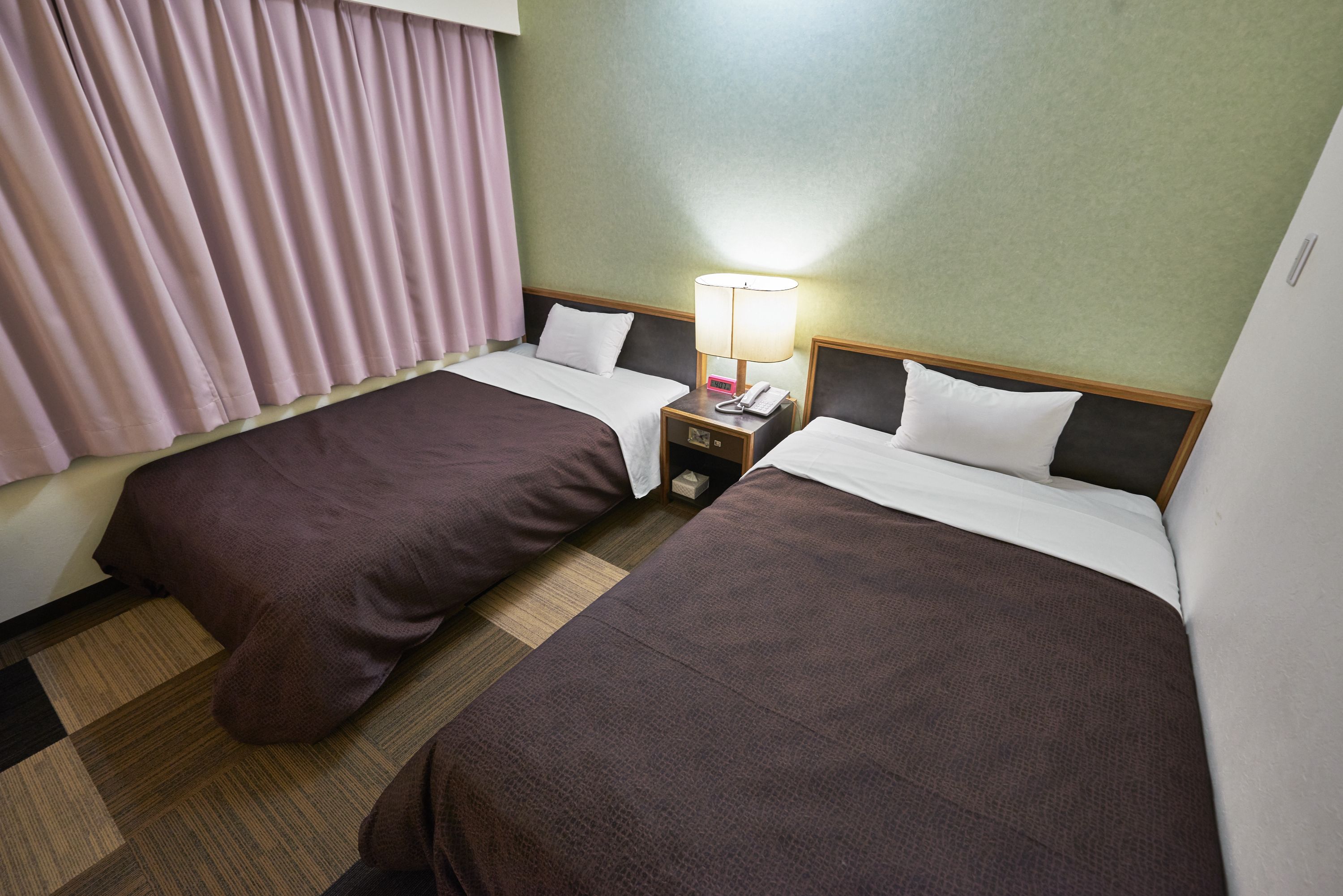 A hotel room with two beds, a nightstand with a lamp, and a window with curtains.