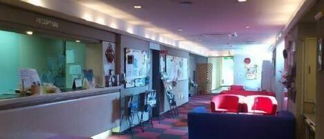 A hotel lobby with a reception desk, a waiting area with red chairs, and a colorful checkered floor.