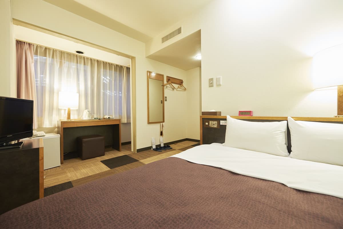 A hotel room with a bed, a desk with a computer, a TV, a mirror, and a window with curtains.
