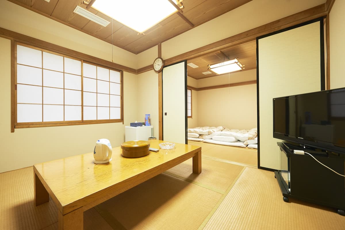 A traditional Japanese room with tatami flooring, a wooden table, a television, and a sliding door leading to a sleeping area.